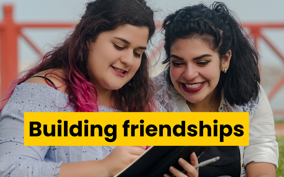 Two young women sitting outdoors. They are smiling at what one is writing in a notebook. Text reads building friendships